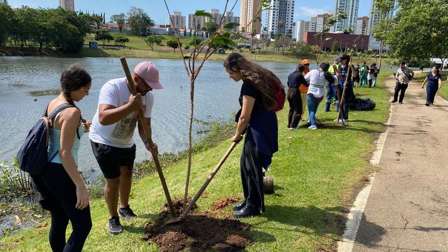 Fotografia em perspectiva ao nível do solo mostrando uma ação coletiva de plantio de árvores no Parque Ecológico, às margens de um lago. Em primeiro plano, do lado esquerdo, três pessoas trabalham na terra ao redor de uma muda recém-plantada. Um homem de camiseta branca, bermuda preta e boné rosa segura uma enxada para acomodar a terra na base do tronco, observado por uma mulher de regata clara e mochila nas costas. Do lado direito da muda, outra mulher de cabelos longos e roupas escuras auxilia