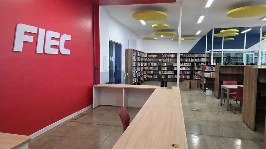 Foto da nova Biblioteca da Fiec. O espaço possui uma parede vermelha com a identificação “Fiec” em letras brancas, mesas e cadeiras de madeira e estantes com livros ao fundo. O ambiente é amplo, iluminado e conta com detalhes decorativos amarelos no teto. Fim da descrição.