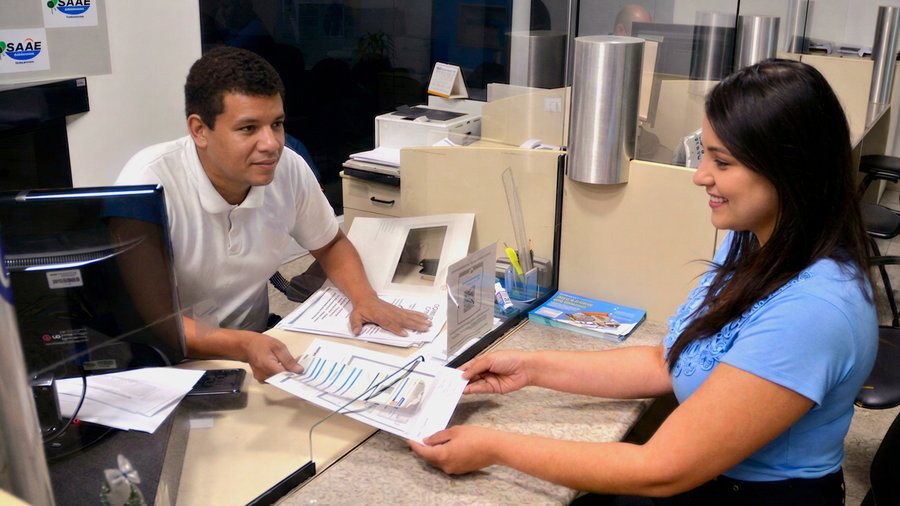 Um homem realizando um atendimento em um balcão de atendimento com um vidro que divide ele de uma mulher. Ele está entregando um papel parecido com uma conta de água. Fim da descrição.