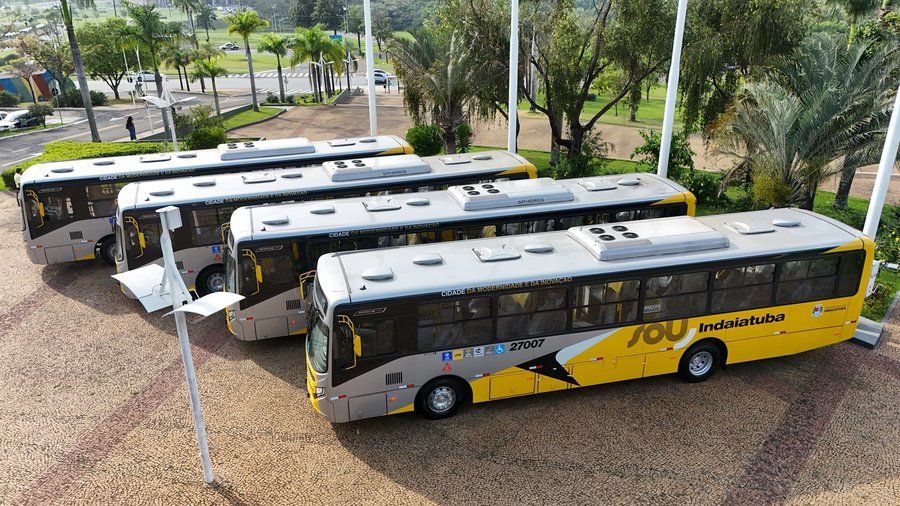 Fotografia em perspectiva aérea e em ângulo diagonal mostrando quatro ônibus novos da frota do transporte coletivo urbano estacionados em sequência sobre uma praça pavimentada em dia ensolarado. Os veículos possuem identidade visual padrão nas cores prata e amarelo, com detalhes em preto. Na lateral do ônibus em primeiro plano, destaca-se o logotipo da operadora "SOU Indaiatuba" em letras garrafais amarelas e pretas, o número de identificação "27007", o brasão do município e pictogramas azuis de