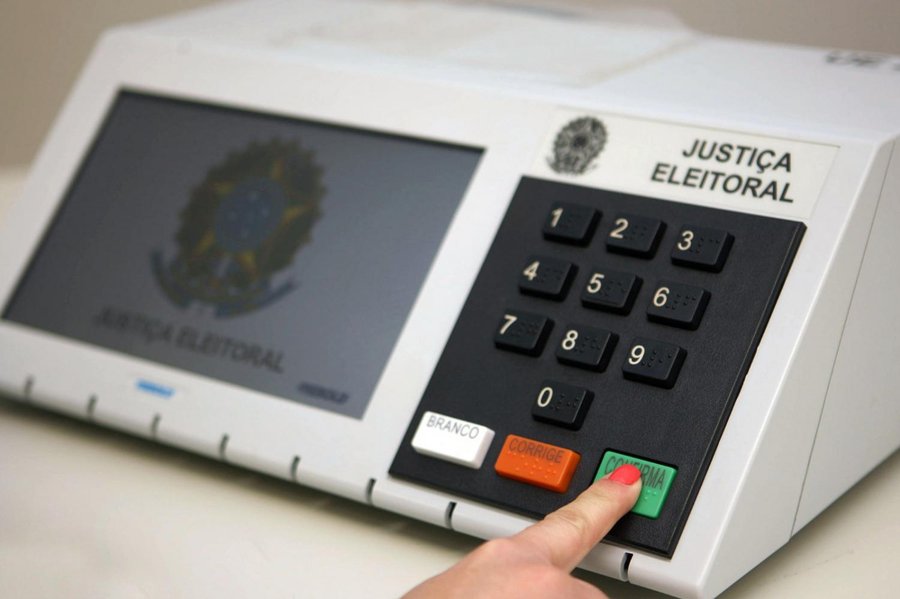a imagem mostra uma urna da Justiça Eleitoral e um dedo de uma mão apertando a tecla confirma, na cor verde. O dedo está com a unha pintada de vermelho.