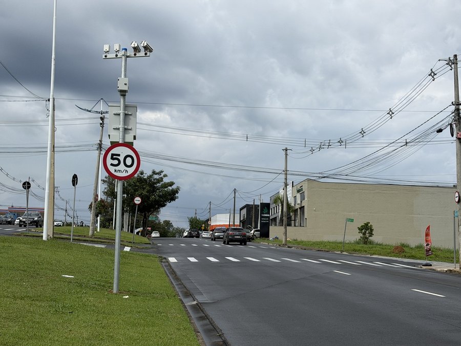 Fotografia em perspectiva ao nível do solo mostrando uma avenida urbana com sinalização de trânsito e fiscalização eletrônica sob céu nublado. Em primeiro plano, do lado esquerdo, destaca-se um poste metálico fixado no canteiro central de grama verde. O poste possui uma placa circular com borda vermelha indicando o limite de velocidade de "50 km/h" em letras e números pretos sobre fundo branco. Acima da placa, no topo do suporte, estão instaladas câmeras e sensores de radar de fiscalização eletr