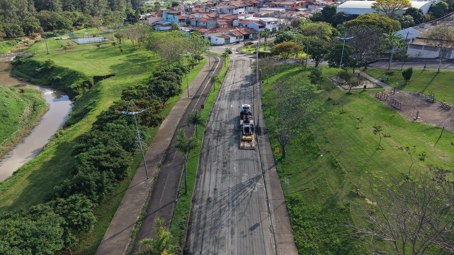 Fotografia aérea capturada por drone mostrando o trecho da Marginal do Parque Ecológico em obras de recapeamento asfáltico, ladeada por áreas verdes em dia ensolarado. No centro da imagem, a pista passa por processo de fresagem e preparação do piso asfáltico, onde é possível ver caminhões e maquinários de pavimentação em operação ao longo da via. Paralela à pista, do lado esquerdo, estende-se uma ciclovia ladeada por arborização e, mais à esquerda, o leito de um rio margeado por gramados e veget