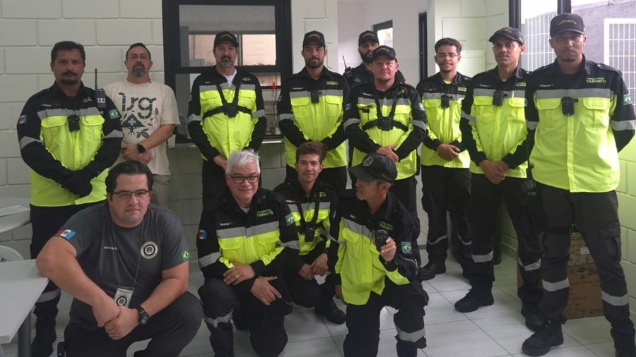Fotografia em ambiente interno mostrando a equipe de agentes de trânsito e transporte de Indaiatuba com câmeras corporais (bodycams). O grupo é composto por doze pessoas dispostas em dois planos. Em primeiro plano, quatro integrantes estão agachados. Ao fundo, oito integrantes estão em pé alinhados lado a lado. A maioria dos agentes veste o uniforme operacional completo da Secretaria de Mobilidade Urbana, composto por jaquetas e camisas em preto e amarelo de alta visibilidade com a palavra "TRÂN
