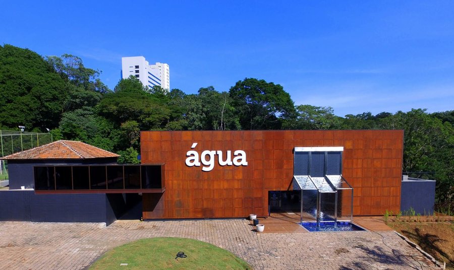 Foto panorâmica do prédio do Museu da Água feita durante o dia, sob um céu azul limpo e sem nuvens. A fachada do prédio tem área envidraçada e painéis de tom ferrugem com a palavra "água" escrita em letras minúsculas e brancas no centro. O edifício é rodeado por vegetação densa e árvores verdes ao fundo. Fim da descrição.