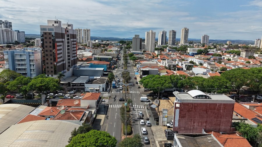 Fotografia aérea capturada por drone mostrando a extensão da Avenida Presidente Kennedy sob céu parcialmente nublado. A via se estende em linha reta pelo centro da imagem, dividida por um canteiro central estreito com arborização e iluminação pública. Em ambas as pistas, observam-se veículos em circulação e faixas de pedestres demarcadas nos cruzamentos. Ao longo de toda a avenida, há um contraste entre edifícios residenciais altos e prédios comerciais modernizados com casas térreas de telhados