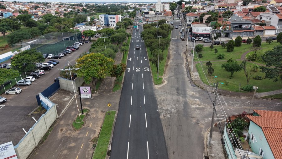 Fotografia aérea capturada por drone exibindo uma ampla via urbana recém pavimentada sob luz natural em dia nublado. No centro da imagem, destaca-se uma pista de asfalto novo e escuro dividida em três faixas de rolamento com pintura horizontal branca recém aplicada. No piso, há a numeração "1", "2" e "3" identificando as faixas, acompanhada por setas que indicam as direções de tráfego (conversão à esquerda, seguir em frente e conversão à direita). Paralela à pista recém recapeada, do lado direit