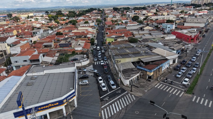 Fotografia aérea capturada por drone mostrando a vista panorâmica de um cruzamento urbano e vias recém-pavimentadas no município sob céu parcialmente nublado. No centro da imagem, destaca-se uma rua com asfalto novo e escuro, onde se observa o fluxo contínuo de veículos e a sinalização horizontal recém-pintada. A via conta com faixa de pedestres zebrada no cruzamento, demarcações de retenção e pictograma indicativo de área de espera para bicicletas/ciclistas no semáforo. À esquerda, em primeiro