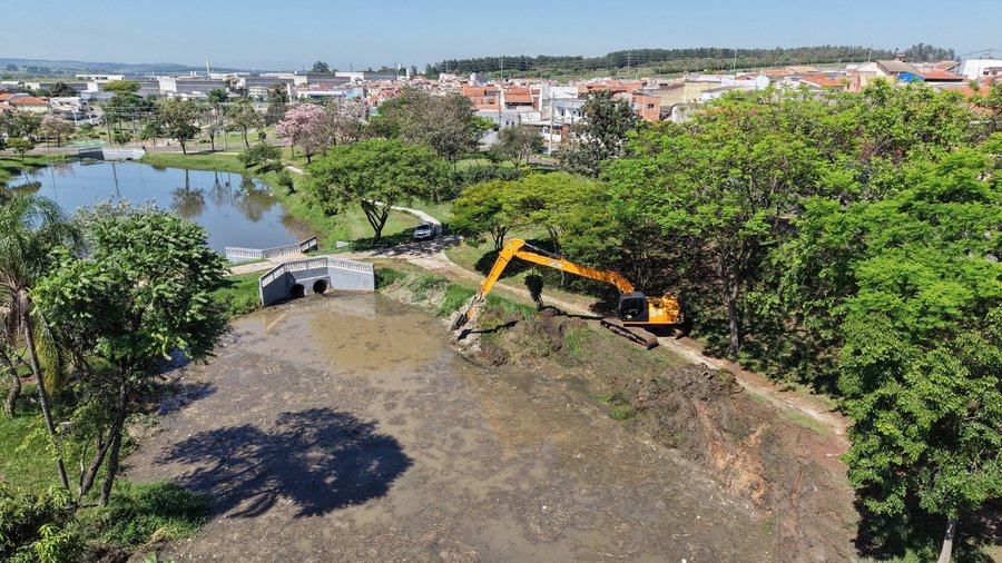 Fotografia aérea capturada por drone mostrando as obras de desassoreamento e manutenção no lago do Jardim Paulista em um dia ensolarado com céu limpo. Em primeiro plano, observa-se o leito parcial do lago rebaixado, exibindo a área com lodo e sedimentos expostos. À direita, uma escavadeira hidráulica de cor amarela com braço longo está posicionada na margem, realizando a remoção da terra e a conformação do talude entre árvores de copa densa e verde. Ao centro, uma pequena estrutura em alvenaria