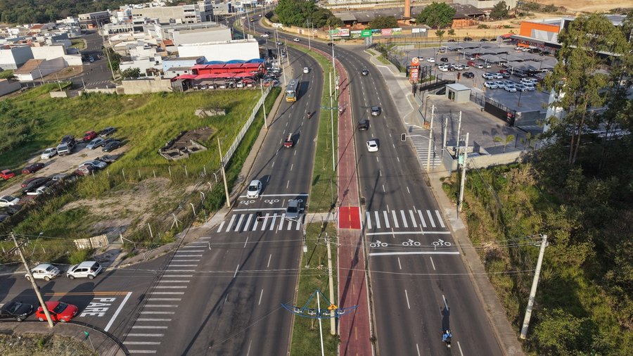Fotografia aérea capturada por drone mostrando a vista em perspectiva da Avenida Manoel Ruz Peres, de pista dupla com canteiro central sob iluminação solar diurna. No centro da imagem, o canteiro gramado é cortado por uma ciclovia pintada na cor vermelha, que possui uma travessia sinalizada em vermelho vivo no cruzamento. Em ambas as pistas, com três faixas de rolamento em cada sentido, há fluxo de carros, motos e um ônibus. O cruzamento em primeiro plano conta com sinalização horizontal complet