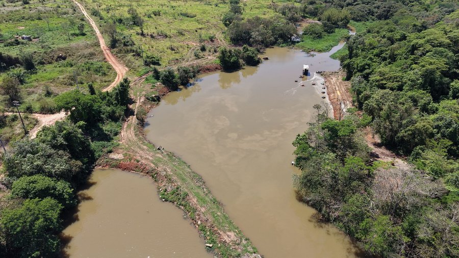 Fotografia aérea do Ribeirão Piraí, mostrando o trecho onde ocorre a captação de água. O corpo hídrico é cercado por vegetação densa e campos verdes. Também aparecem estradas de terra batida contornando o curso d'água, além de pequenas edificações ao redor da margem. Fim da descrição.
