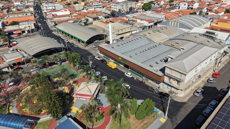 Fotografia aérea capturada por drone exibindo a vista panorâmica de uma área urbana residencial e comercial sob céu aberto. Em primeiro plano, no canto inferior esquerdo, destaca-se uma praça pública totalmente revitalizada, composta por calçamentos coloridos em círculos vermelhos, amarelos e cinzas, área de playground, canteiros com palmeiras e arborização densa. Ao centro, uma rua com asfalto novo, escuro e liso cruza a imagem na diagonal. Ao longo dessa via recém-pavimentada, observa-se o trâ