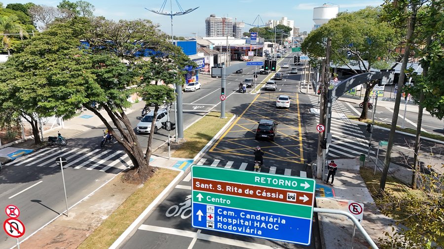 Fotografia aérea capturada por drone mostrando a vista em perspectiva do cruzamento e fluxo de veículos na Avenida Presidente Vargas sob iluminação solar diurna. Em primeiro plano, no canto inferior central e direito, destaca-se uma grande placa indicativa de trânsito em pórtico metálico com orientações nas cores verde, marrom e azul. O cruzamento possui sinalização horizontal completa, incluindo uma ampla caixa amarela de marcação de área confluente (não feche o cruzamento) no centro da pista,