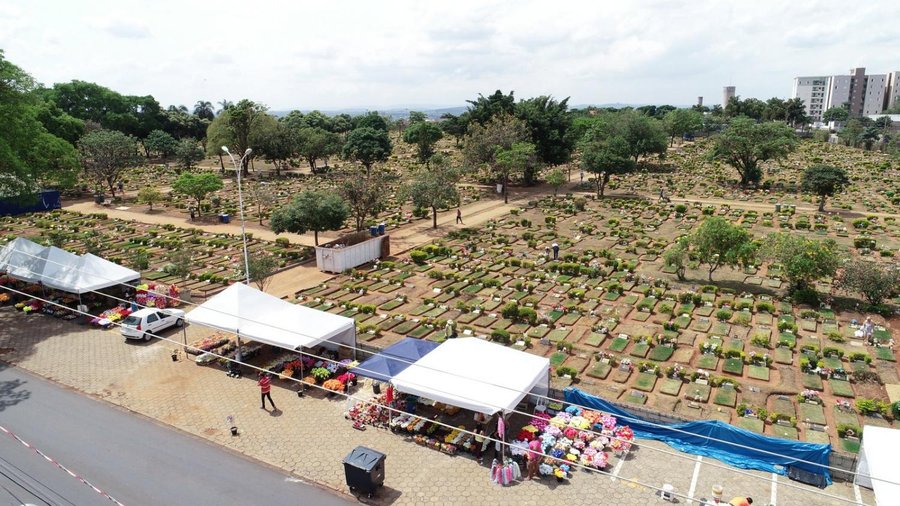 A foto colorida aérea mostra um cemitério do modelo jardim, com túmulos separados por caminhos de terra, bastante arborizado, com tendas brancas de comércio de flores posicionadas na calçada.