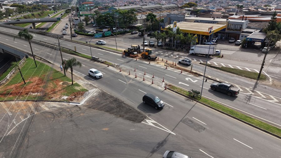 Fotografia aérea capturada por drone exibindo obras de infraestrutura e intervenção viária em uma via de pista dupla sob iluminação solar diurna. Ao centro da imagem, em uma das faixas de rolamento bloqueada temporariamente por cones de sinalização laranjas e brancos, observam-se duas máquinas amarela de infraestrutura pesada realizando trabalhos no pavimento. O trânsito de veículos como carros e picapes segue pelas faixas liberadas ao lado. À esquerda, no plano superior, destaca-se a elevação d