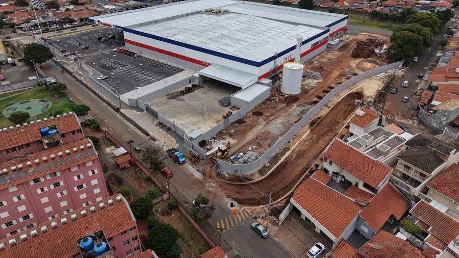 Fotografia aérea capturada por drone mostrando as obras de terraplenagem para a abertura de uma nova via ao lado de um grande centro comercial sob iluminação diurna. No centro e na parte superior da imagem, destaca-se o amplo galpão da loja Paulistão Atacadista, com fachada nas cores azul, branca e vermelha, além de seu estacionamento privativo à esquerda e docas de carga e descarga na área posterior. Ladeando o complexo comercial à direita, estende-se uma ampla faixa em curva de terra vermelha